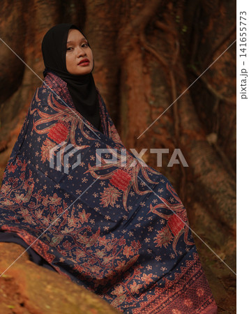 Portrait of a beautiful young Muslim woman wearing an elegant black hijab and traditional red Jambi Batik, posing confidently in a rustic industrial warehouse, modern modest fashion concept. Portrait of a beautiful young Muslim woman wearing an elegant black hijab and traditional red Jambi Batik, posing confidently in a rustic industrial warehouse, modern modest fashion concept. 141655773