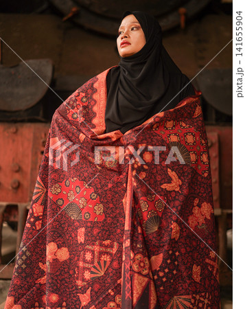Beautiful Indonesian Muslim woman wearing a black hijab and traditional red Jambi Batik dress, posing confidently in an industrial warehouse setting. 141655904