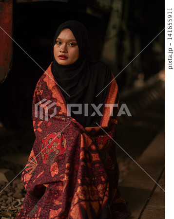 Beautiful Indonesian Muslim woman wearing a black hijab and traditional red Jambi Batik dress, posing confidently in an industrial warehouse setting. Beautiful Indonesian Muslim woman wearing a black hijab and traditional red Jambi Batik dress, posing confidently in an industrial warehouse setting. 141655911