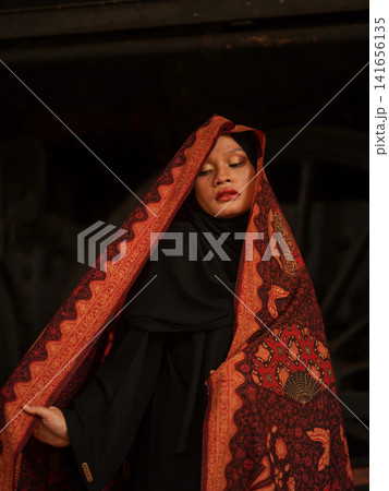 Fashionable young Asian Muslimah in traditional red Jambi Batik and dark hijab standing in an abandoned industrial space, showcasing modern modest style. Fashionable young Asian Muslimah in traditional red Jambi Batik and dark hijab standing in an abandoned industrial space, showcasing modern modest style. 141656135