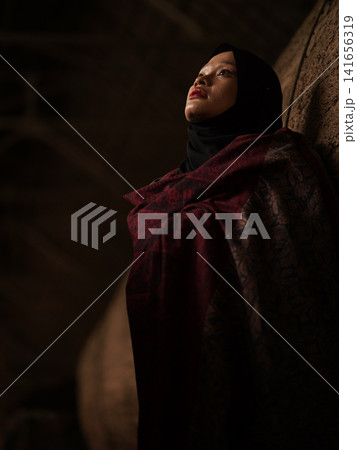 Confident young Asian woman showcasing modest fashion in a red Jambi Batik dress and black hijab, posing in a rustic warehouse environment. 141656319