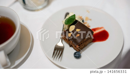 A delicious slice of chocolate cake topped with almonds, cream, mint, and a blueberry, served on a white plate next to a cup of tea, perfect for a gourmet dessert. A delicious slice of chocolate cake topped with almonds, cream, mint, and a blueberry, served on a white plate next to a cup of tea, perfect for a gourmet dessert. 141656331