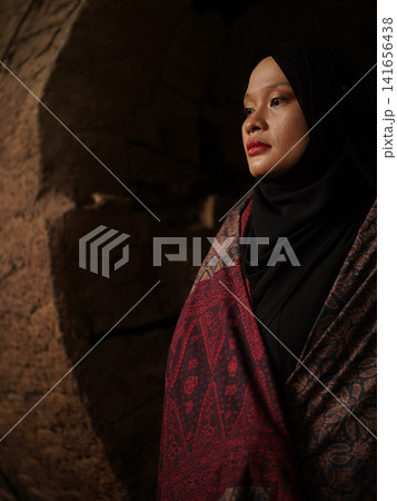 Confident young Asian woman showcasing modest fashion in a red Jambi Batik dress and black hijab, posing in a rustic warehouse environment. 141656438