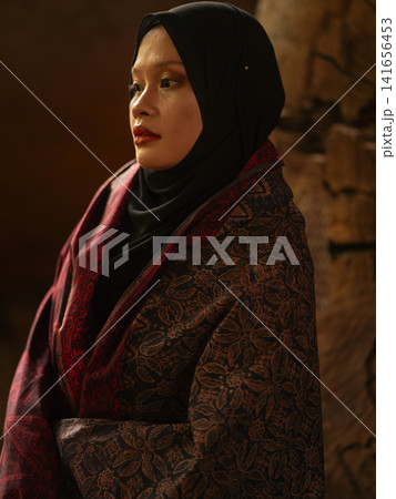 Confident young Asian woman showcasing modest fashion in a red Jambi Batik dress and black hijab, posing in a rustic warehouse environment. 141656453