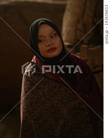 A fashionable young woman wearing a traditional black hijab and an elegant long red dress standing confidently against a weathered, vintage green door. Perfect for modest fashion, cultural, and lifest 141656613