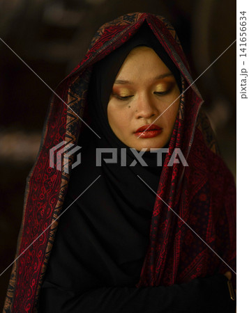 A fashionable young woman wearing a traditional black hijab and an elegant long red dress standing confidently against a weathered, vintage green door. Perfect for modest fashion, cultural, and lifest A fashionable young woman wearing a traditional black hijab and an elegant long red dress standing confidently against a weathered, vintage green door. Perfect for modest fashion, cultural, and lifest 141656634
