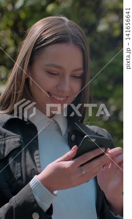 Vertical video. Young woman enjoying city streets, chatting with friends, using cellphone outdoors, sunset background. 141656641