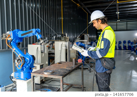 Engineer in high-visibility gear checks blue robotic arm in modern factory workshop setting 141657706