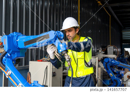 Engineer in high-visibility vest adjusts blue robotic arm in industrial workshop with corrugated metal walls 141657707