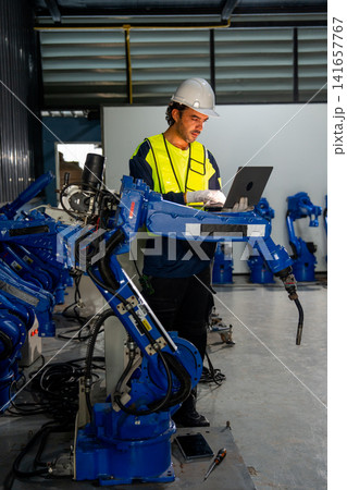 Engineer in safety gear programs blue robotic arm with laptop in industrial workshop with corrugated metal walls 141657767