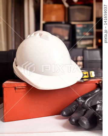 White hard hat resting on a red metal toolbox, with work gloves and a measuring tape, depicting labor, construction, and safety for manual workers and skilled trades 141659037