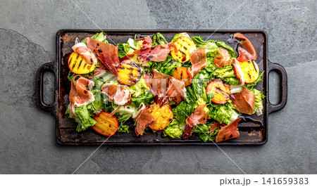Grilled peach and prosciutto salad with green lettuce, balsamic glaze and wine on dark background 141659383