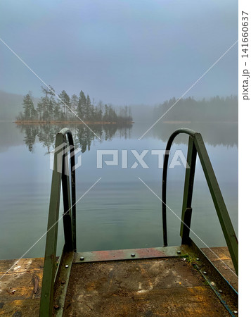 Misty lake with wooden dock and ladder reflecting pine island Misty lake with wooden dock and ladder reflecting pine island 141660637
