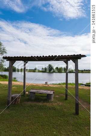 Rustic riverside shelter with corrugated roof and bench by calm river Rustic riverside shelter with corrugated roof and bench by calm river 141661515