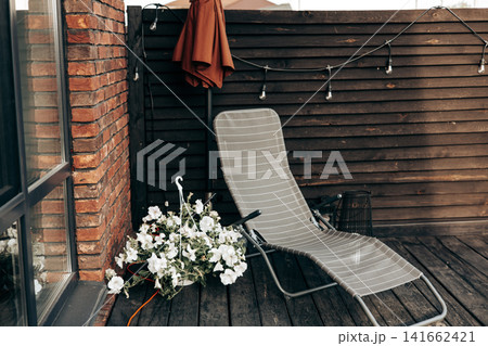 Modern outdoor terrace or patio with lounge chair, blooming white flowers and wooden privacy fence. Cozy backyard relaxation space at a contemporary home, showcasing comfort and stylish living. Modern outdoor terrace or patio with lounge chair, blooming white flowers and wooden privacy fence. Cozy backyard relaxation space at a contemporary home, showcasing comfort and stylish living. 141662421