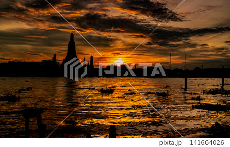 Silhouette of Wat Arun at dramatic sunset over the Chao Phraya River in Bangkok, Thailand, glowing reflections, clouds and iconic Buddhist temple architecture. Famous historic Buddhist landmark. 141664026