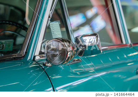 A polished chrome bullet-style side mirror and round turn signal light are mounted at the base of the windshield pillar on a beautifully restored teal classic American automobile. The bright chrome 141664464