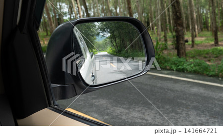 View through a car side mirror showing an empty asphalt road. Lined with green trees during a daytime drive. Blurry of a motorized tricycle is visible at the far end of the road. View through a car side mirror showing an empty asphalt road. Lined with green trees during a daytime drive. Blurry of a motorized tricycle is visible at the far end of the road. 141664721