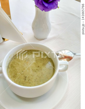 Creamy green vegetable soup served in white bowl with spoon and flower vase on table 141665766