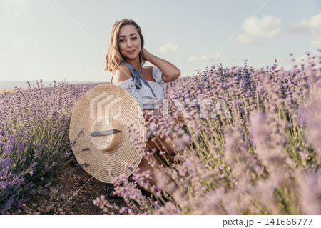 Woman lavender field holding a large straw hat while sitting among blooming flowers in the countryside at sunset 141666777
