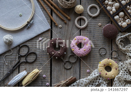 Handmade knitting and sewing accessories with colorful donut pincushions lace embroidery hoop scissors and yarn on rustic wooden background top view craft composition Handmade knitting and sewing accessories with colorful donut pincushions lace embroidery hoop scissors and yarn on rustic wooden background top view craft composition 141667297