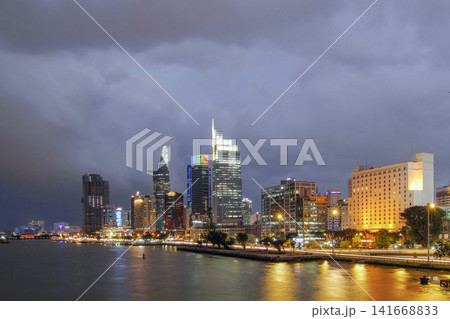 Wide nighttime cityscape showing illuminated modern towers, riverbank buildings, calm reflections on water, and dramatic storm clouds above busy downtown lights along waterfront, with skyline, city Wide nighttime cityscape showing illuminated modern towers, riverbank buildings, calm reflections on water, and dramatic storm clouds above busy downtown lights along waterfront, with skyline, city 141668833