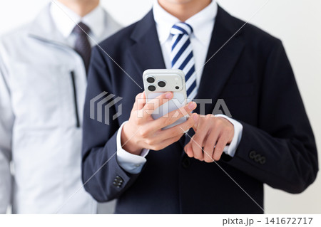 スマホを操作するリクルートスーツを着た若い男性と作業服のミドル男性 スマホを操作するリクルートスーツを着た若い男性と作業服のミドル男性 141672717