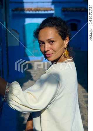 Woman portrait smiling in a sunlit blue-street wearing gold earrings and a white blouse Woman portrait smiling in a sunlit blue-street wearing gold earrings and a white blouse 141672734
