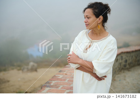 Contemplative woman in white dress overlooking a foggy hillside from a brick wall Contemplative woman in white dress overlooking a foggy hillside from a brick wall 141673182