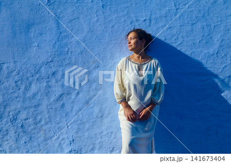Woman in white dress leaning against a blue wall bathed in warm sunlight and long shadow 141673404