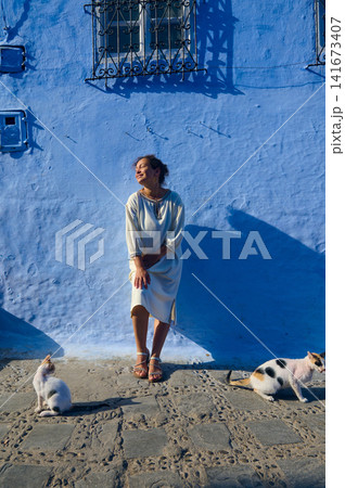 Woman relaxing against bright blue wall with cats on sunny cobblestone street in Morocco Woman relaxing against bright blue wall with cats on sunny cobblestone street in Morocco 141673407