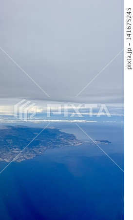 Aerial view of island coastline surrounded by deep blue ocean and layered clouds seen from airplane. Natural seascape with land, water and sky, travel and tourism concept, calm atmosphere, freedom and Aerial view of island coastline surrounded by deep blue ocean and layered clouds seen from airplane. Natural seascape with land, water and sky, travel and tourism concept, calm atmosphere, freedom and 141675245