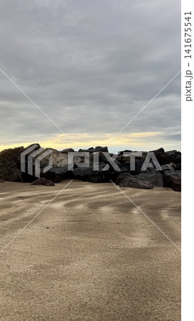 Natural rocky coastline with rippled sand patterns in foreground and dark volcanic stones under dramatic cloudy sunset sky, minimal seascape background for travel, nature, mindfulness, freedom and cal Natural rocky coastline with rippled sand patterns in foreground and dark volcanic stones under dramatic cloudy sunset sky, minimal seascape background for travel, nature, mindfulness, freedom and cal 141675541
