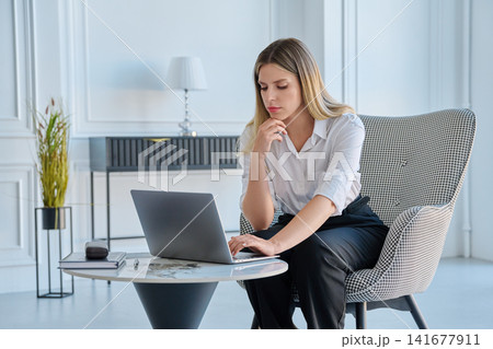 Young focused businesswoman working on laptop at small table in modern office Young focused businesswoman working on laptop at small table in modern office 141677911