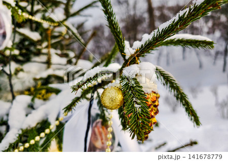 A tree stands in the snow decorated with ornaments and a pine cone. It is wintertime and the landscape is covered in white snow 141678779