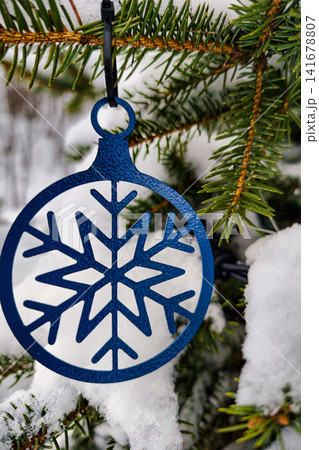 A snowflake ornament is suspended from an evergreen tree branch covered with white snow. The scene shows a natural winter landscape during the holiday time A snowflake ornament is suspended from an evergreen tree branch covered with white snow. The scene shows a natural winter landscape during the holiday time 141678807