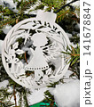 A white ornament shaped like an angel hangs on a Christmas tree. Snow covers the ground and tree, creating a festive atmosphere for the holiday season 141678847