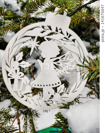 A white ornament shaped like an angel hangs on a Christmas tree. Snow covers the ground and tree, creating a festive atmosphere for the holiday season A white ornament shaped like an angel hangs on a Christmas tree. Snow covers the ground and tree, creating a festive atmosphere for the holiday season 141678847
