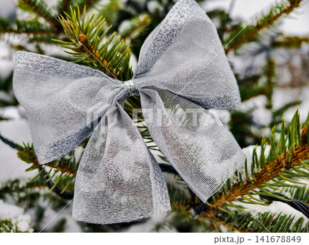A Christmas tree stands decorated with a silver bow and snow-covered branches. The scene shows the festive spirit of the holiday season in winter A Christmas tree stands decorated with a silver bow and snow-covered branches. The scene shows the festive spirit of the holiday season in winter 141678849