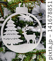 A round ornament shows a tree and a deer. The ornament hangs on a green branch of a decorated tree in a cold winter atmosphere during the holidays 141678851