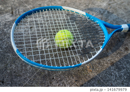 Blue tennis racket with green ball resting in strings on rough gray outdoor asphalt court, close-up sports equipment detail for training and recreational play, with tennis, racket, ball, sport detail 141679979