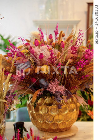 Elegant arrangement of dried flowers, preserved botanicals and ornamental grasses displayed in a decorative golden vase inside a flower shop.  141680333