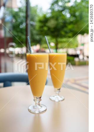 Two fresh banana smoothies served in tall glasses with paper straws on a table inside a cozy cafeteria. Creamy tropical beverages against a softly blurred street view through the cafe window Two fresh banana smoothies served in tall glasses with paper straws on a table inside a cozy cafeteria. Creamy tropical beverages against a softly blurred street view through the cafe window 141680370