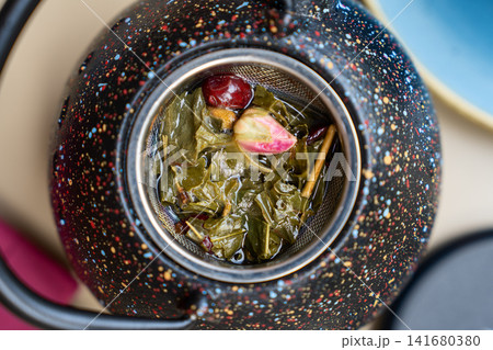 Fragrant botanical tea blend with dried foliage, colorful fruits and blooming rosebud inside a metal strainer within a speckled porcelain kettle Fragrant botanical tea blend with dried foliage, colorful fruits and blooming rosebud inside a metal strainer within a speckled porcelain kettle 141680380