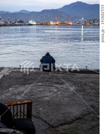 lonely man sits on the pier in the evening. A man is sitting in the port area. Loneliness concept. Port city in winter. lonely man sits on the pier in the evening. A man is sitting in the port area. Loneliness concept. Port city in winter. 141682555