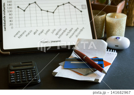 Business workspace representing financial planning, data analysis, and productivity with a tablet displaying a graph, a calculator, credit cards, a pen, and receipts on a dark surface Business workspace representing financial planning, data analysis, and productivity with a tablet displaying a graph, a calculator, credit cards, a pen, and receipts on a dark surface 141682967