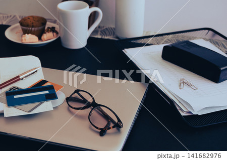 Desk showing a laptop. Eyeglasses. Multiple credit cards. A pen. And a stack of business documents. Creating a scene of financial planning. Busy work Desk showing a laptop. Eyeglasses. Multiple credit cards. A pen. And a stack of business documents. Creating a scene of financial planning. Busy work 141682976