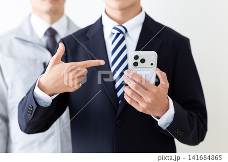 スマホを指差すリクルートスーツを着た若い男性と作業服のミドル男性 スマホを指差すリクルートスーツを着た若い男性と作業服のミドル男性 141684865