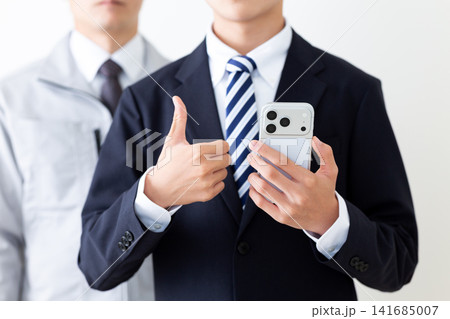 スマホを持って親指を立てるリクルートスーツを着た若い男性と作業服のミドル男性 スマホを持って親指を立てるリクルートスーツを着た若い男性と作業服のミドル男性 141685007