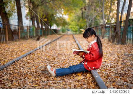 Asian girl reading book on railway track in autumn park, focused emotion, education and peaceful outdoor learning concept Asian girl reading book on railway track in autumn park, focused emotion, education and peaceful outdoor learning concept 141685565